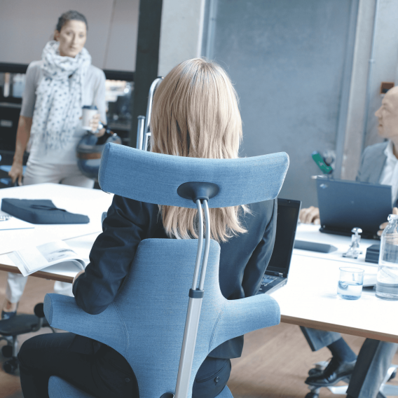 Detailansicht eines blauen HAG Capisco 8107 von der Rueckseite mit einer Frau drauf sitzend im Setting eines Meetings