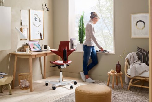 Capisco Puls 8010 als Stuhl im kreativen Home-Office Setting mit einer Frau am Fenster stehend in einem Buch blätternd